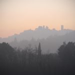 Silhouette di una città collinare con torri e alberi, vista attraverso la foschia e la nebbia all'alba o al tramonto, con un cielo grigio e arancione sullo sfondo.