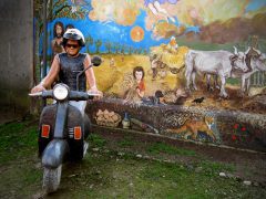 Una persona con casco e occhiali da sole guida uno scooter nero davanti a un murale colorato che raffigura la vita rurale, tra persone, animali, un Orto e un paesaggio vivace, perfetto per i fan dei nuovi arrivati.