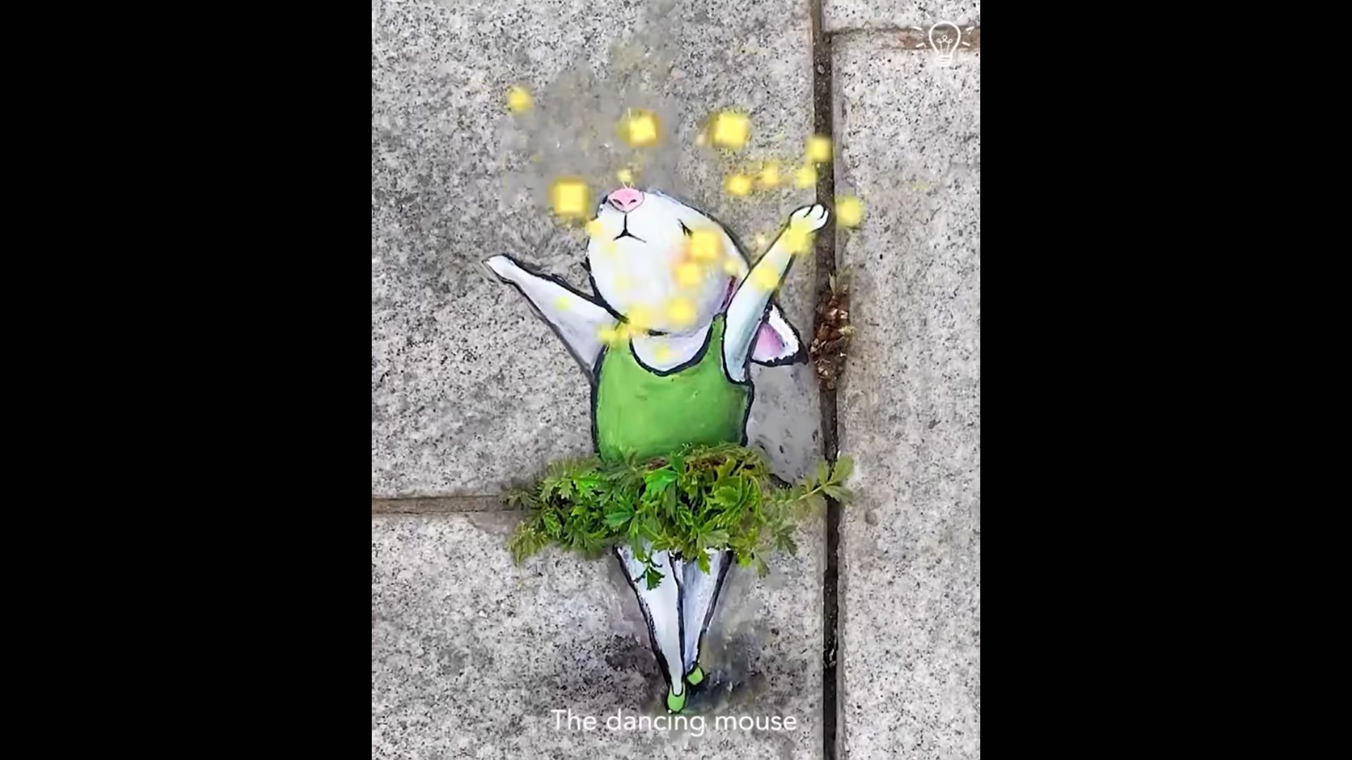 Un disegno di un topo con un vestito verde e un tutù fatto di muschio vero si trova sulla pavimentazione. Il topo sta in punta di piedi con le braccia alzate, circondato da quadrati gialli, e la didascalia recita: Il topo ballerino.