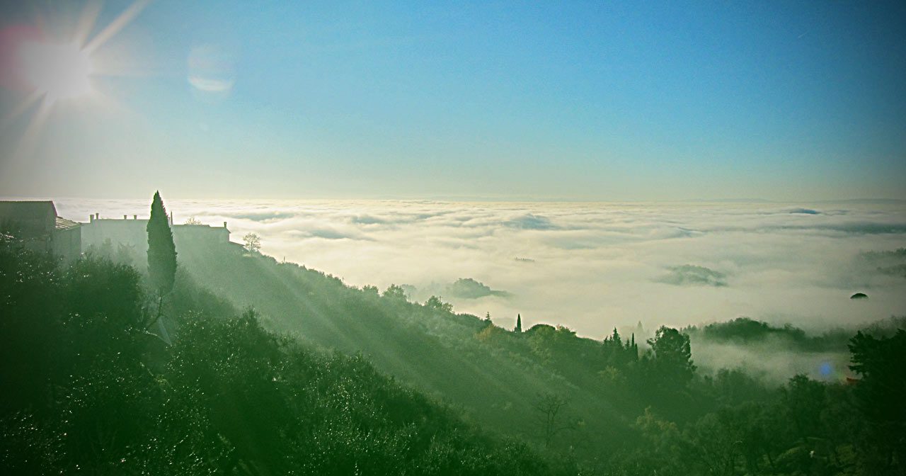 La luce del sole illumina una collina nebbiosa coperta di alberi; una coltre di nuvole si estende sul paesaggio sotto un cielo azzurro e limpido, con sagome di edifici e alti cipressi in primo piano.